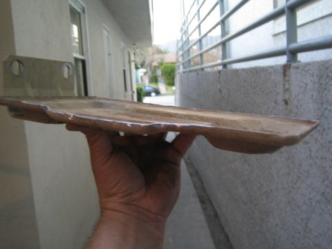 1990 Ford Bronco - metal battery tray held edge-on, showing its wavy welded lip, against a stucco wall and metal railing.