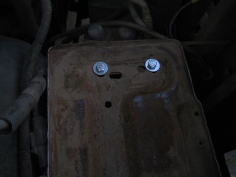 1990 Ford Bronco - rusty metal battery tray secured with two shiny new bolts, mounted under a vehicle amid wiring and components.