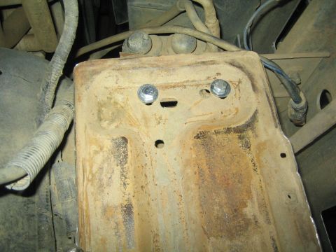 1990 Ford Bronco - heavily rusted battery tray bolted into a vehicle's engine compartment, surrounded by corrugated wiring and hoses.