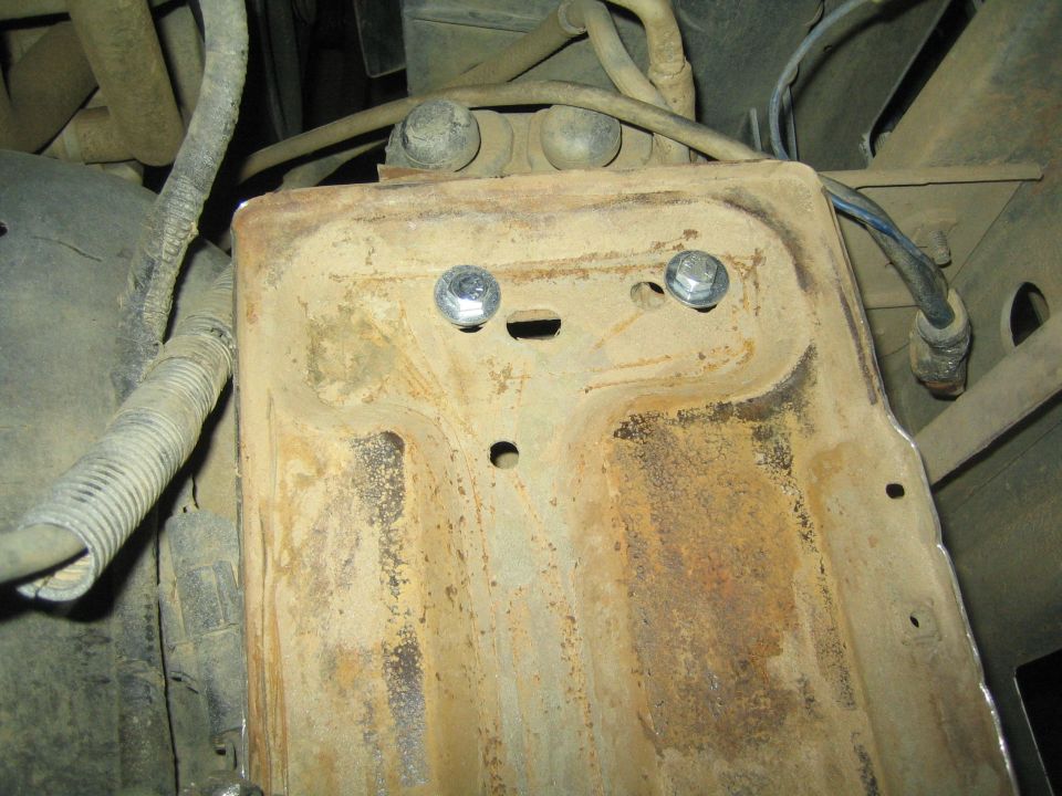 Rusty battery tray bolted in place under a vehicle with surrounding wiring and hoses