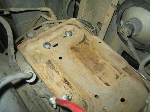1990 Ford Bronco - corroded battery tray bolted under a vehicle, with a red cable clamp and electrical connector visible nearby.