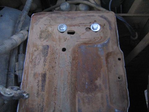 1990 Ford Bronco - rust-stained metal battery tray fastened with two bolts and a slotted hole inside an engine compartment.