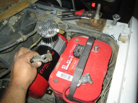 1990 Ford Bronco - hand positioning a small ExtremeAire air compressor next to a red automotive battery in a cluttered engine bay.