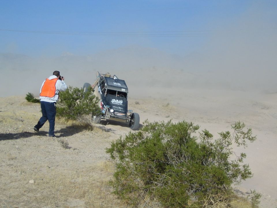 Photographer in orange vest shooting a dark off-road race buggy in desert dust