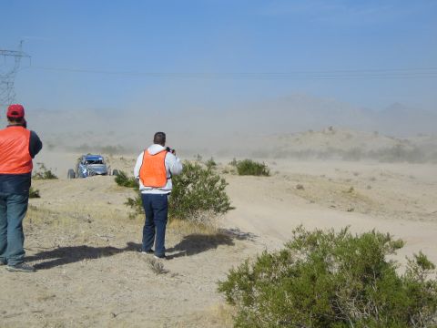 Two men in orange vests watch a blue off-road buggy raise dust under power lines