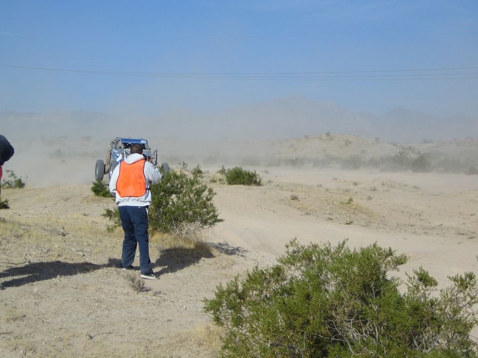 Person in orange vest photographing a blue off-road buggy in the dusty desert