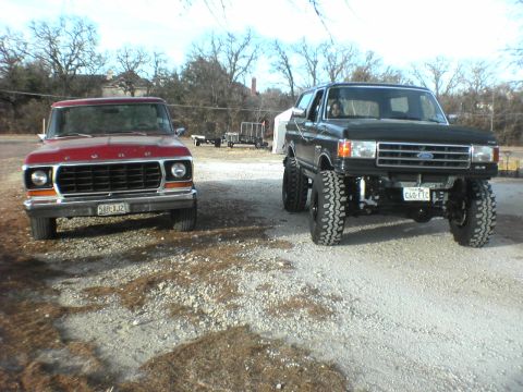 1991 Ford Bronco - Two Ford trucks, a lifted Bronco and a red pickup, are parked outside on a gravel lot.