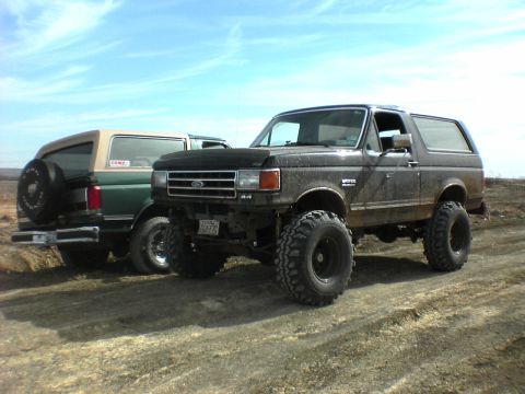 1991 Ford Bronco - Two modified Ford Broncos sit side-by-side on a dusty, off-road path under a bright blue sky.