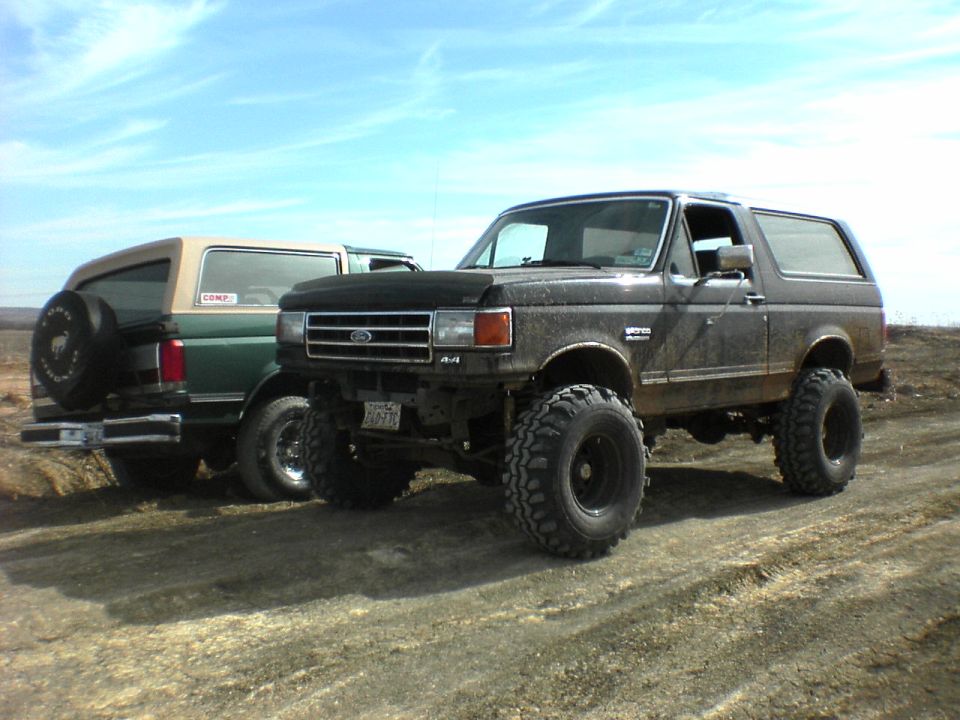 Two lifted Ford Broncos parked on a dirt road. The front one is dark colored, the rear one is green with a tan top