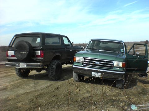 1991 Ford Bronco - Two Ford trucks, a green F-150 and a black Bronco, are parked on a dirt incline under a partly cloudy sky.