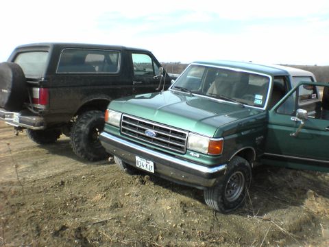 1991 Ford Bronco - A black 1991 Ford Bronco and a green Ford pickup truck are parked side-by-side on a dirt lot.