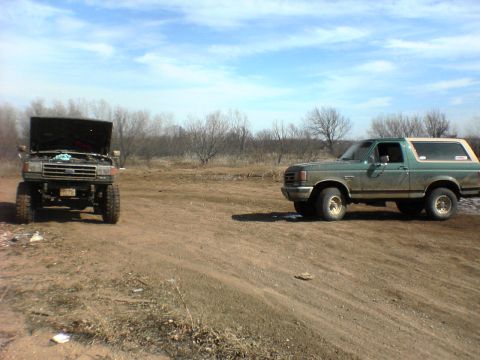 1991 Ford Bronco - Two Broncos sit on a dirt lot under a partly cloudy sky, with one having its hood open.