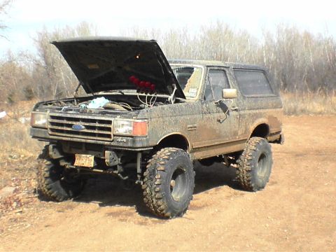 1991 Ford Bronco - This lifted Ford Bronco is covered in mud, with its hood open, parked on a dirt road.