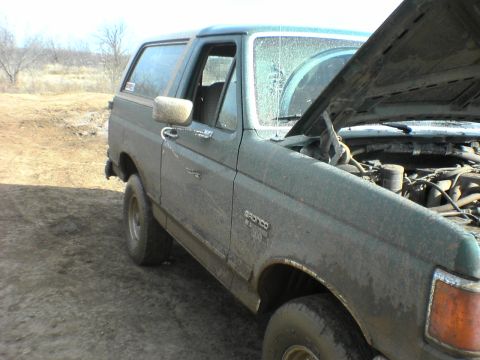 1991 Ford Bronco - A muddy 1991 Ford Bronco sits on a dirt road with its hood open, revealing the engine compartment.