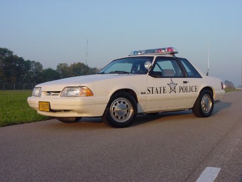 1993 Ford Mustang - A white 1993 Ford Mustang police car is parked on the side of a road under a clear sky.