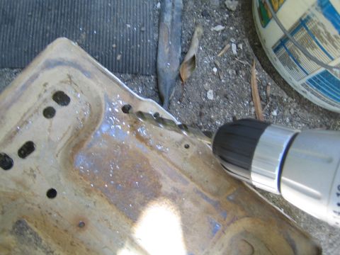 1990 Ford Bronco - cordless drill bores holes into a rust-stained stamped sheet-metal tray that has been wetted down, lying on concrete outdoors.