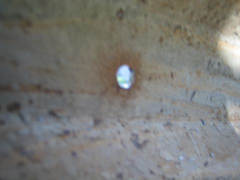 1990 Ford Bronco - Blurry macro shot of a single small drilled hole in a pitted, rust-brown sheet-metal surface with light showing through.