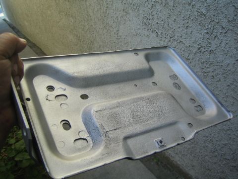 1990 Ford Bronco - hand holds up a freshly bare, contoured stamped sheet-metal battery-tray panel with mounting holes, against a stucco wall.