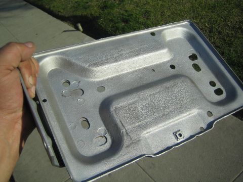 1990 Ford Bronco - hand holds up a bare galvanized stamped sheet-metal tray with multiple mounting holes outdoors over a sidewalk and grass.
