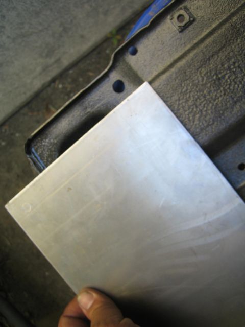 1990 Ford Bronco - hand holds a flat rectangular brushed sheet-metal blank above a dark blue-black stamped panel resting on the ground.