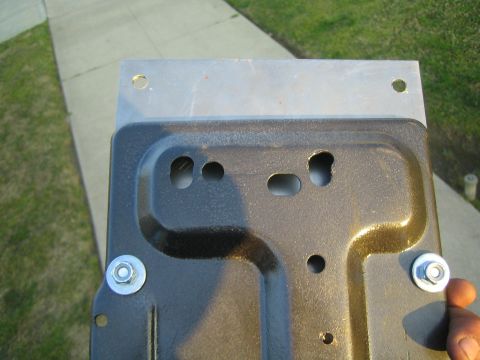 1990 Ford Bronco - hand holds a black-coated stamped metal tray fastened with zinc bolts to a flat plate, viewed from the back over pavement.