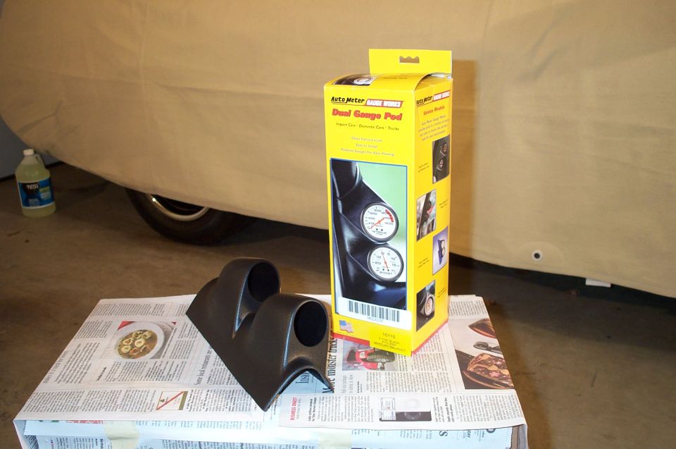 Black dual A-pillar gauge pod and yellow Auto Meter box on newspaper in a garage