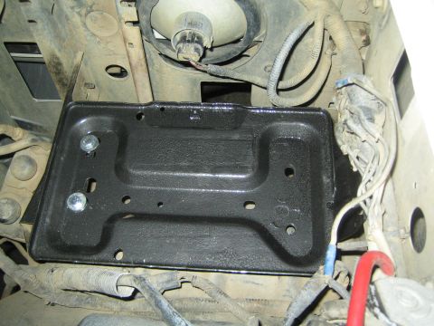 1990 Ford Bronco - black-coated stamped battery tray sits mounted in an engine bay surrounded by wiring harnesses and a red battery cable.