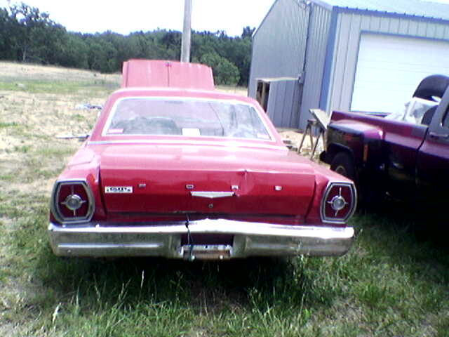 Rear view of red 1965 Ford Galaxie 500 on grass with hood raised, maroon pickup beside it