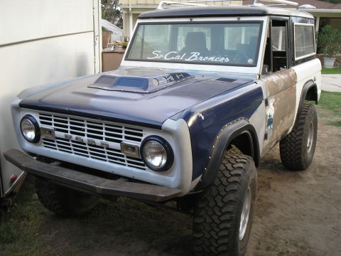 1966 Ford Bronco - Three-quarter front view of a lifted early Ford Bronco with a dark blue hood featuring a cowl scoop, silver-primered front fenders, weather…