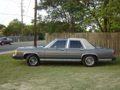 1988 Ford LTD Crown Victoria - A gray 1988 Ford Crown Victoria LTD is seen from its side, parked on a grassy area next to a wooden fence.