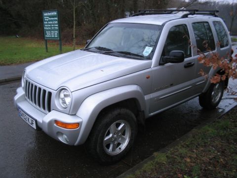 1992 Ford Bronco - A silver Jeep Liberty SUV is parked on a wet road next to a grassy area with a sign in the background.