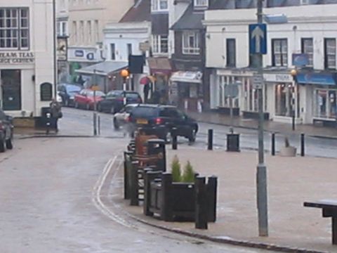 1992 Ford Bronco - A dark SUV drives down a wet street lined with shops on a cloudy day.
