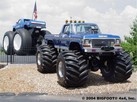 1979 Ford Bronco - A blue 1979 Ford Bronco monster truck, branded Bigfoot, is parked next to another monster truck under a cloudy sky.