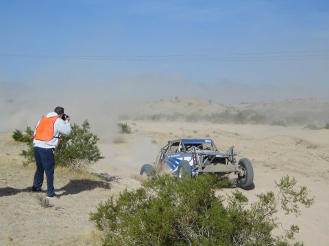 Photographer in orange vest shooting a blue off-road buggy kicking up dust in the desert