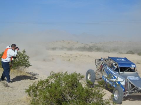 Photographer in orange vest beside a blue numbered off-road buggy raising dust