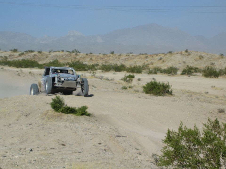 Silver tube-frame off-road buggy catching air over a desert berm