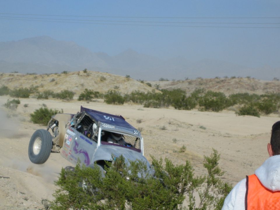 Purple off-road buggy launching off a berm as a spectator in orange vest watches