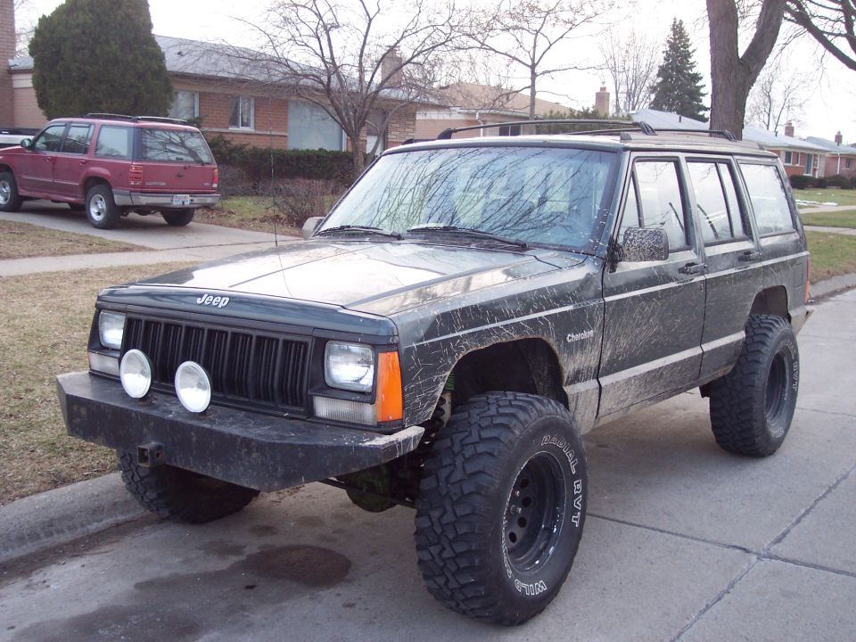 Dark green 1993 Jeep Cherokee with aftermarket bumper and large tires, parked on a residential street