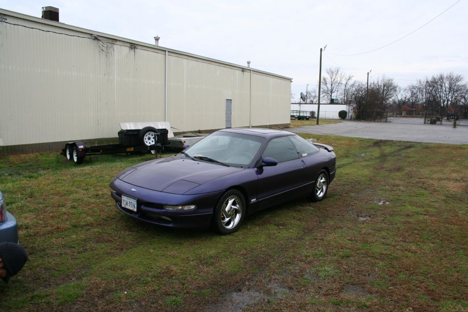 Purple Ford hatchback coupe on polished wheels parked on grass by a metal building