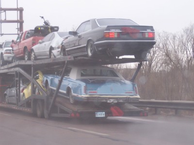 1972 Ford Ranchero - A car carrier truck transports several vehicles, including a blue sedan on the lower deck and a black coupe on the upper deck.