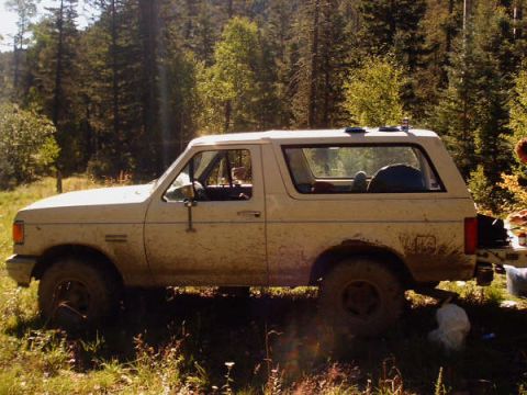 1991 Ford Bronco - A muddy 1991 Ford Bronco sits off-road with trees in the background.