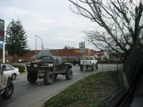 1985 Ford F-150 - A heavily modified pickup truck with oversized tires and a custom roll cage is seen from the rear, driving on a street with other vehicles.