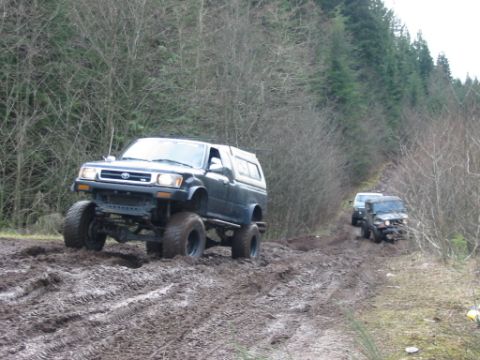 1985 Ford F-150 - Two off-road vehicles navigate a muddy trail through a dense forest with bare trees.
