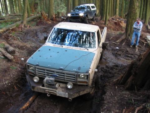 1985 Ford F-150 - Two trucks navigate a challenging muddy path in a dense forest, with one vehicle deeply mired.