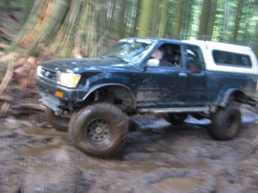 Dark blue pickup truck with white camper shell driving off-road through mud and trees
