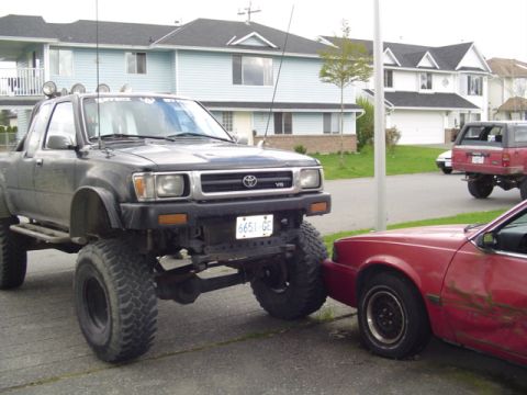 1985 Ford F-150 - A heavily modified black Toyota pickup truck with oversized tires is parked close to a red sedan, with other vehicles and houses in the bac…