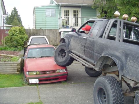 1985 Ford F-150 - A black pickup truck is parked on top of a red sedan, with a white cap on the sedan's bed.