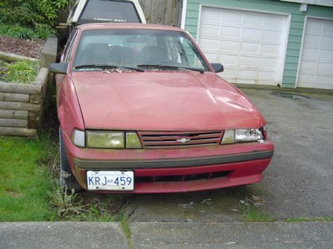 1985 Ford F-150 - A red sedan with a dented front end and a broken headlight sits parked in front of a garage.