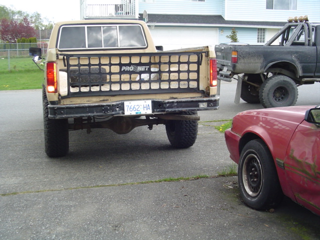 Rear view of a tan Ford F-series truck with a Pro Net tailgate insert. A black lifted truck and a red car are also visible