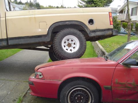1985 Ford F-150 - A tan pickup truck is precariously balanced on top of a red car.